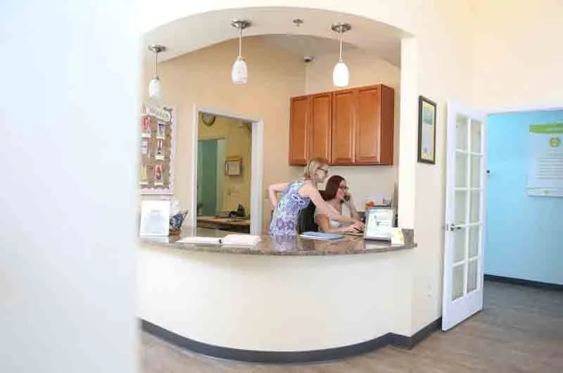 Two women working at a reception desk in a brightly lit office or clinic with arched opening and wooden cabinets.