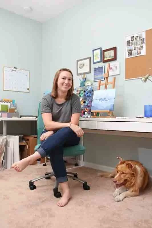 A woman sitting cross-legged on a green office chair in a home office, smiling, with a dog lying on the carpet floor next to her. The room has light blue walls decorated with framed pictures and a bulletin board.