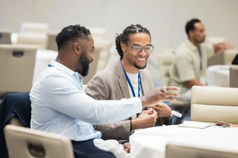 Two men laughing and chatting at a conference table in a professional setting, with another man in the background.