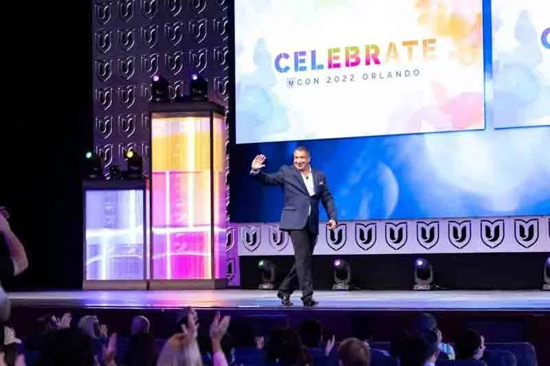 A man in a suit walking on stage at a conference, waving to the audience. Behind him, a screen displays the words "Celebrate ICON 2022 Orlando". The stage has colorful lighting and decorative panels, and the audience is clapping.