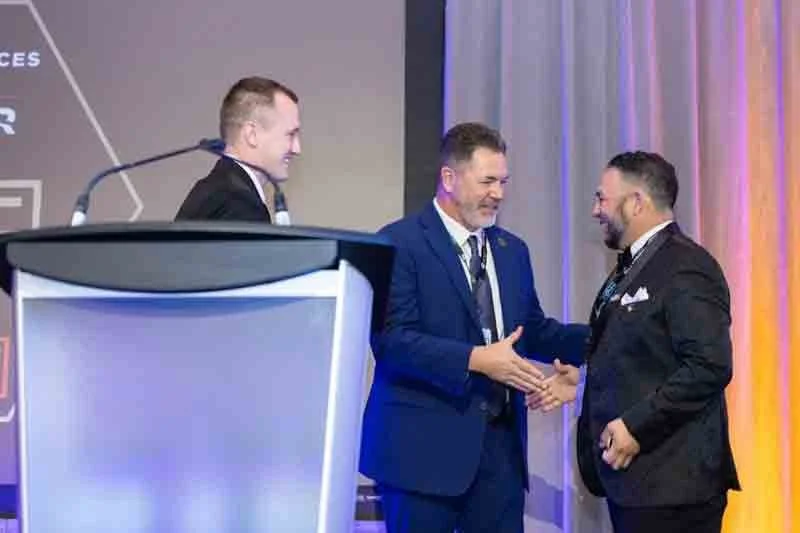 Two men shaking hands on stage at a formal event, with a third man smiling nearby, and subtitles on a screen in the background.