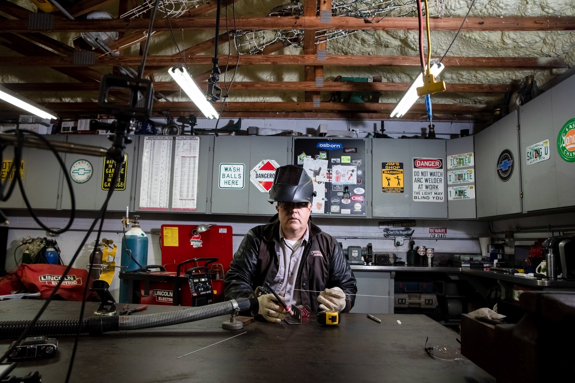 Man working in a garage welding with safety helmet on, surrounded by tools, warning signs, and a cluttered workbench.