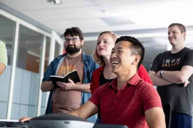 Group of four people in an office, some standing and one sitting, smiling and looking at a computer.