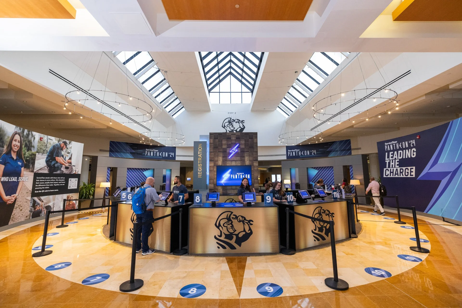 Registration desk at a conference with staff and attendees, large banners with slogans, high ceiling with skylights, and a floor marked with numbered spots for queueing.