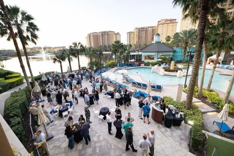 People gathered at an outdoor poolside venue by a river, with high-rise buildings and palm trees in the background, during daytime.