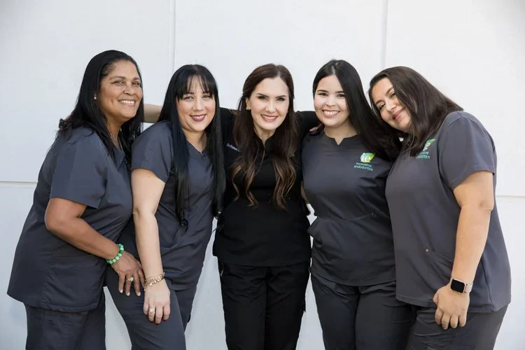 Five women standing close together in scrubs, smiling for a group photo against a plain white wall.