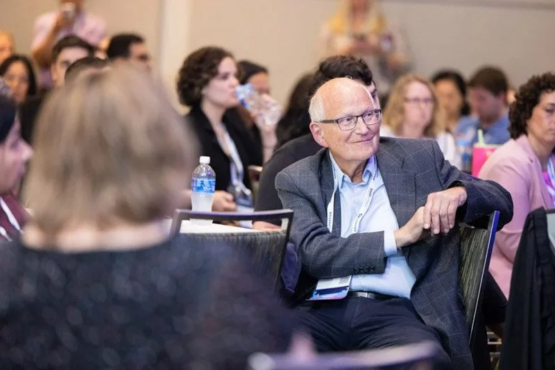 An elderly man with glasses, wearing a gray blazer, seated at a conference or seminar, surrounded by other attendees. Some are drinking water, and there are bottles and notebooks on the tables.