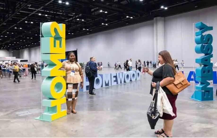 People at a convention with large colorful signs spelling out 'HOME', 'HOMETOWN', and 'HAVE A' inside a spacious indoor venue.