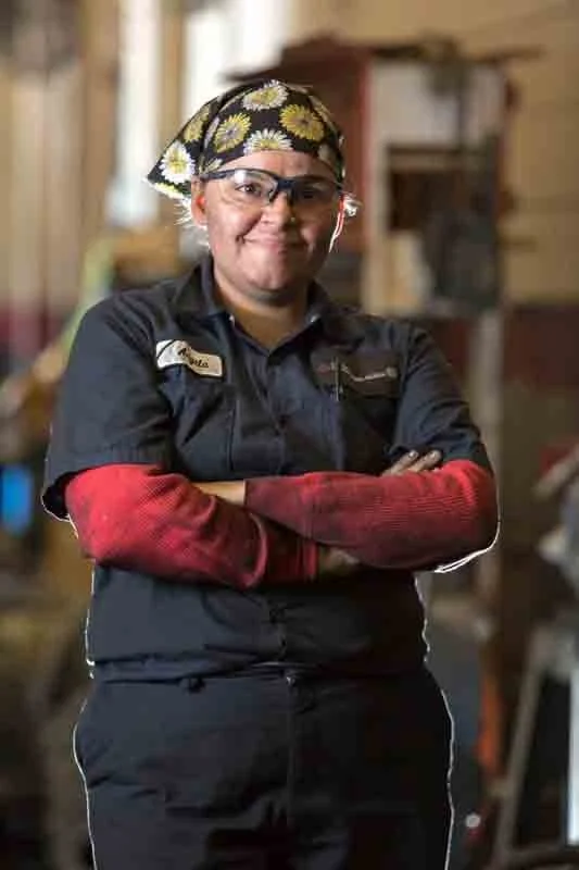 A woman wearing glasses, a bandana with yellow sunflowers, a black work uniform, and red sleeves, standing with arms crossed inside a workshop or garage.