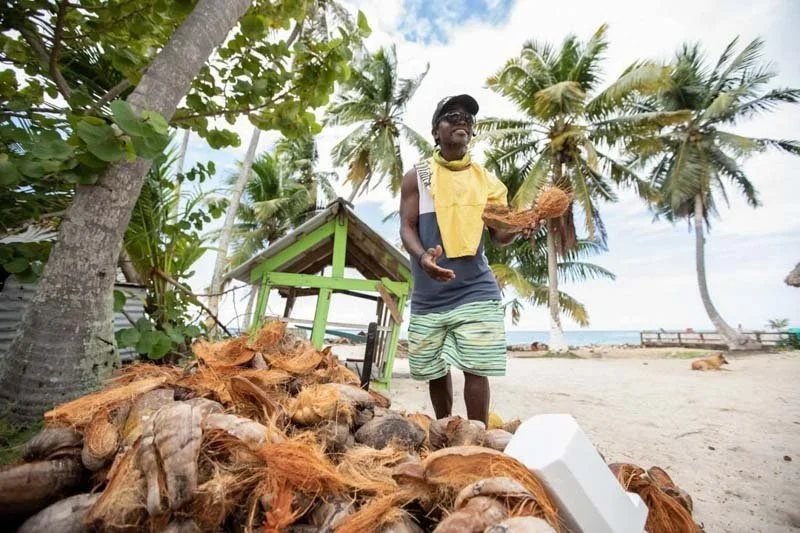 Man standing on a sandy beach among coconuts, palm trees, and a small green hut, holding a coconut shell, with the ocean and a cloudy sky in the background.