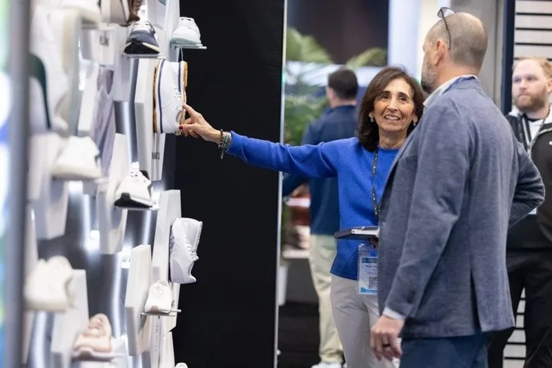 A woman and a man are at a shoe display, with the woman pointing at a white shoe on the wall. The woman is smiling and wearing a blue top, and the man is wearing a gray jacket. Others are in the background at what appears to be a trade show or exhibi
