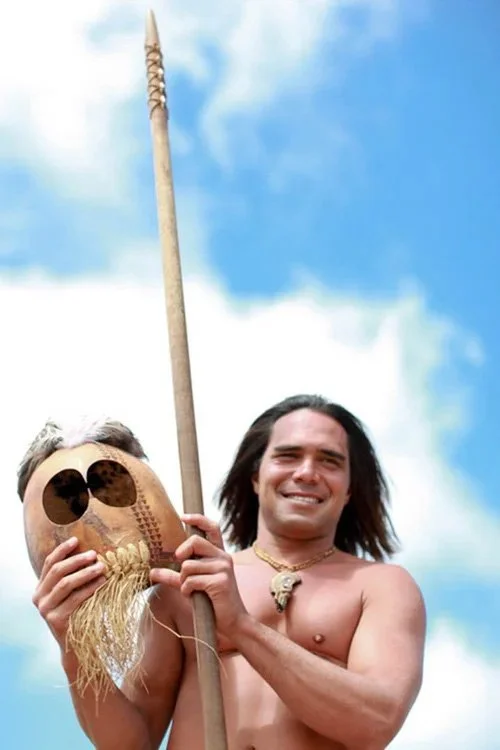 A shirtless man with long dark hair is smiling and holding a tribal spear and a carved wooden mask with sunglasses on it against a blue sky with clouds.