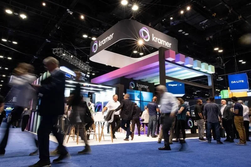 Trade show booth for Kramer with people walking and interacting, large screens displayed, and a modern design with colorful lighting.