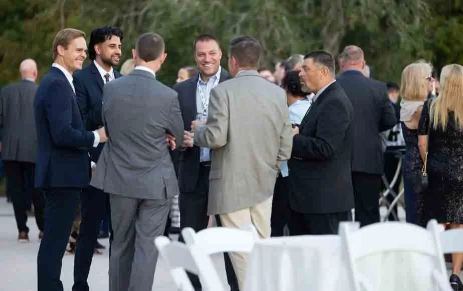 Group of people in formal attire socializing at an outdoor event, with some standing around a table and others holding drinks, in a lush green setting.