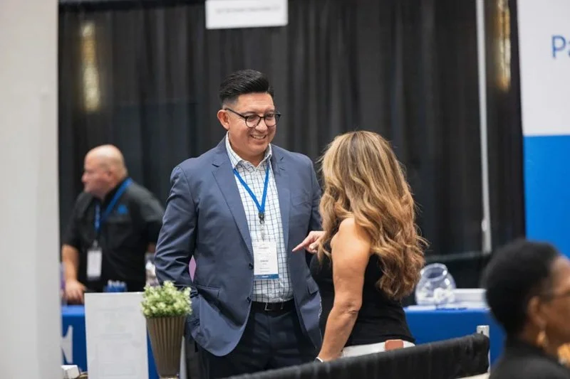 Two people, a man and a woman, are conversing at a conference or exhibition. The man is wearing glasses, a blue suit, and a name badge. The woman has long wavy hair and is dressed in black. There are other attendees and informational booths in the ba