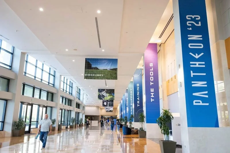 Interior of a spacious lobby with large windows, potted plants, and blue banners displaying conference or event information.