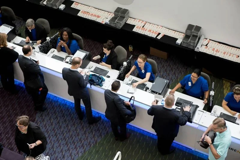 Conference registration or check-in area with staff at a long counter assisting attendees, some using laptops, and others filling out forms; people waiting in line or engaging with staff.