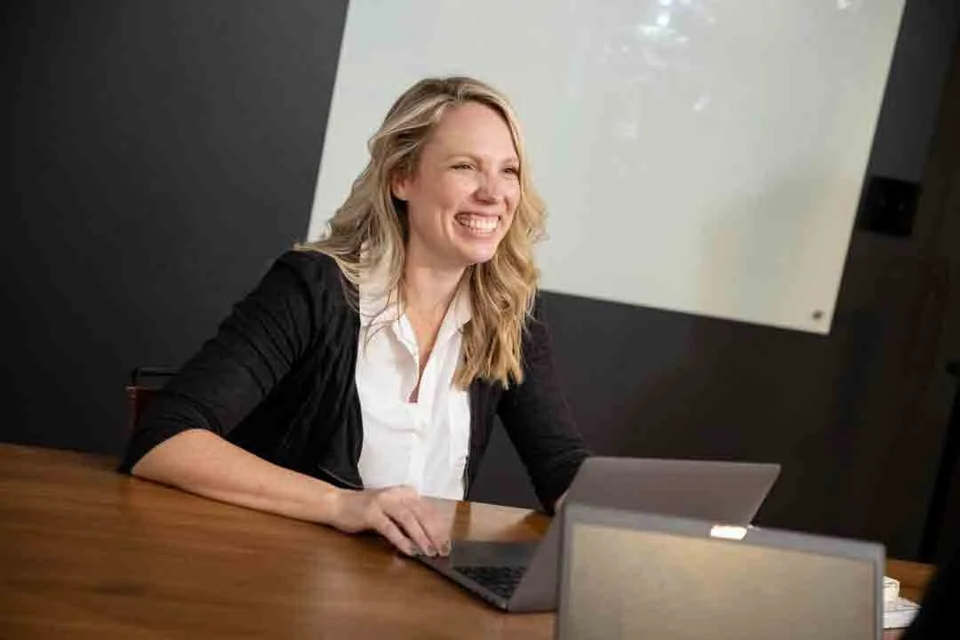 A woman with blond hair is smiling while sitting at a wooden table with a laptop in front of her, in a modern office setting.