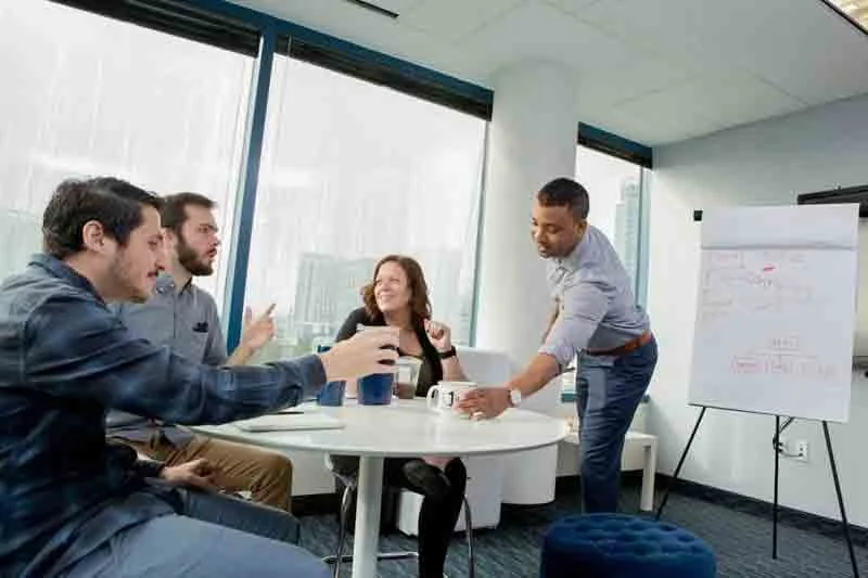 Four people in a modern office space having a business meeting, with large windows and a whiteboard in the background.