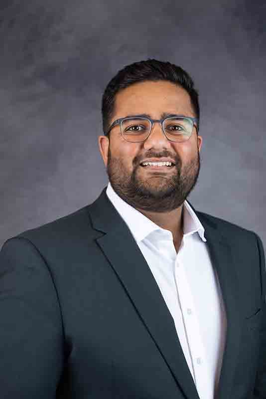 Professional portrait of a man wearing glasses, a white shirt, and a dark suit, smiling against a gray background.