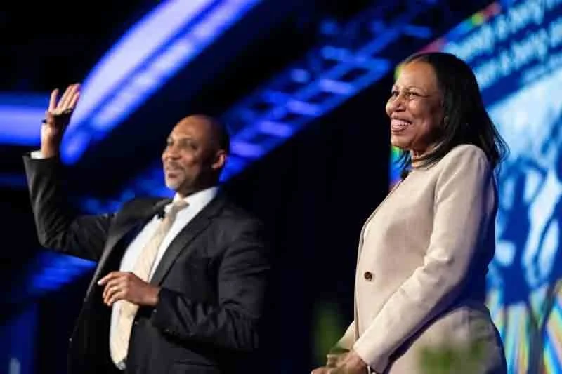 A man and a woman on stage during a formal event, with the man waving and the woman smiling, in front of a bright, colorful background.