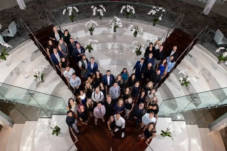Group of people standing on a circular staircase with glass railings, decorated with orchids and flowers, taken from above.