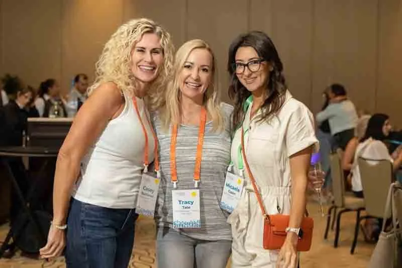 Three women smiling at a conference, wearing name tags and casual attire, standing close together in a large room with other attendees in the background.