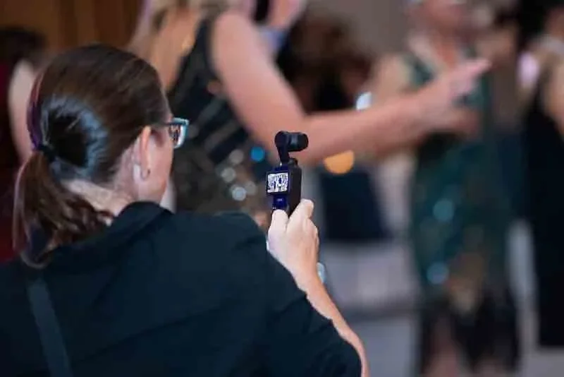 A woman with glasses and brown hair tied back, holding a small video camera, in a crowded setting with people using smartphones.