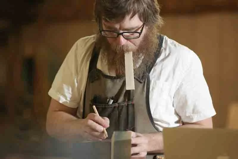 A man with glasses and a beard, wearing a white shirt and an apron, appears to be balancing a rectangular object in his mouth while writing or drawing on a piece of paper.