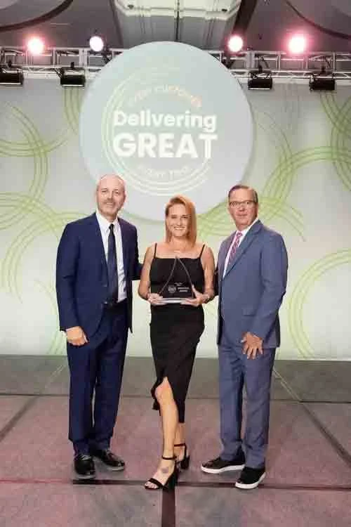 Three people in formal attire standing on a stage, holding an award, with a large screen behind them displaying the words 'Delivering GREAT'.