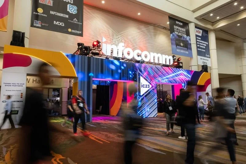 Indoor trade show or conference with a large illuminated display reading 'Infocomm' and a stage with vibrant lights, people walking and networking.