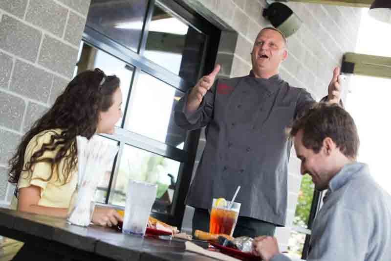 A chef is speaking to two customers at a restaurant table, gesturing with his hands, while the customers listen and smile, with drinks and food on the table.