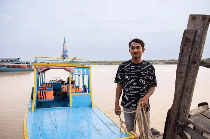 Man standing on a dock near a colorful boat on a river with a child in the boat and boats in the background