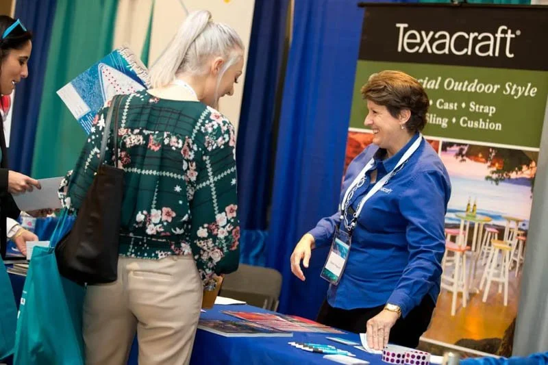 A woman in a blue shirt is smiling and talking to two women at a trade show booth for Texacraft outdoor furniture. The booth has a banner and a table with promotional materials.