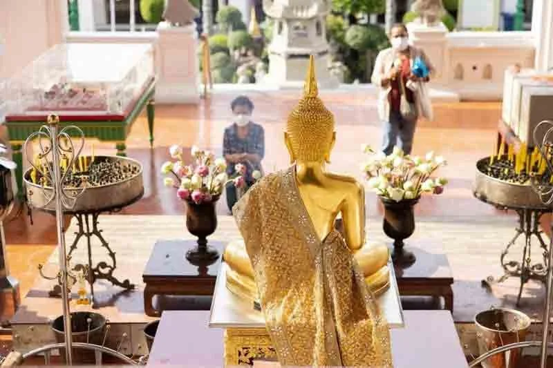 A golden Buddha statue facing away, draped in a cloth, in a temple with offerings of flowers and incense, with visitors observing in the background.