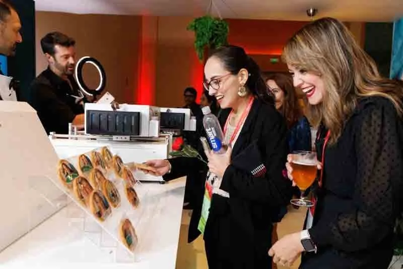 Two women at a food stand smiling and holding drinks, one woman with a water bottle, and the other with a glass of beer. They are in a lively indoor setting with a bartender in the background and colorful lighting.