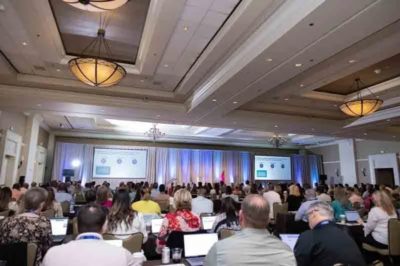 Large conference room filled with attendees sitting at tables, facing a stage with large screens displaying presentations, decorated with curtains and chandeliers overhead.