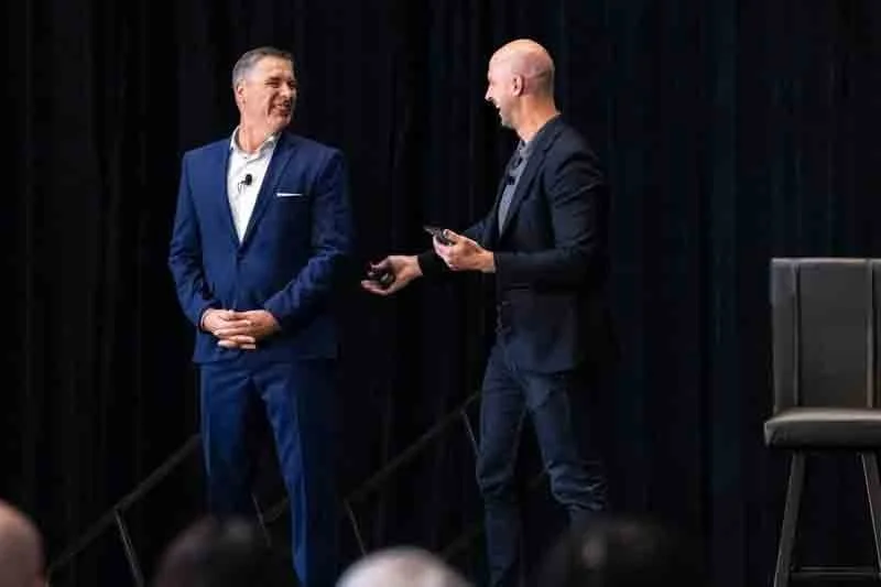 Two men on stage engaged in conversation, one in a blue suit smiling, the other in a dark blazer holding a tablet, with a black curtain backdrop and a solo chair on the stage.