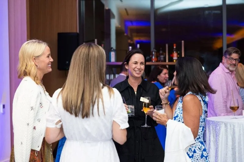 A group of women socializing at a party, holding drinks and engaging in conversations, with a bar in the background.
