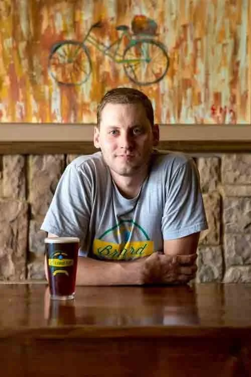 A man sitting at a wooden table with a pint of beer in front of him, in a room with a stone wall and a colorful abstract painting with a bicycle hanging on it.