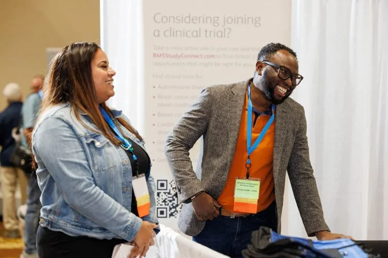 Two smiling conference attendees, a woman and a man, stand at a booth with a white backdrop and informational poster behind them. The woman has long brown hair, wearing a denim jacket and a conference badge, and the man has short hair, glasses, gray 
