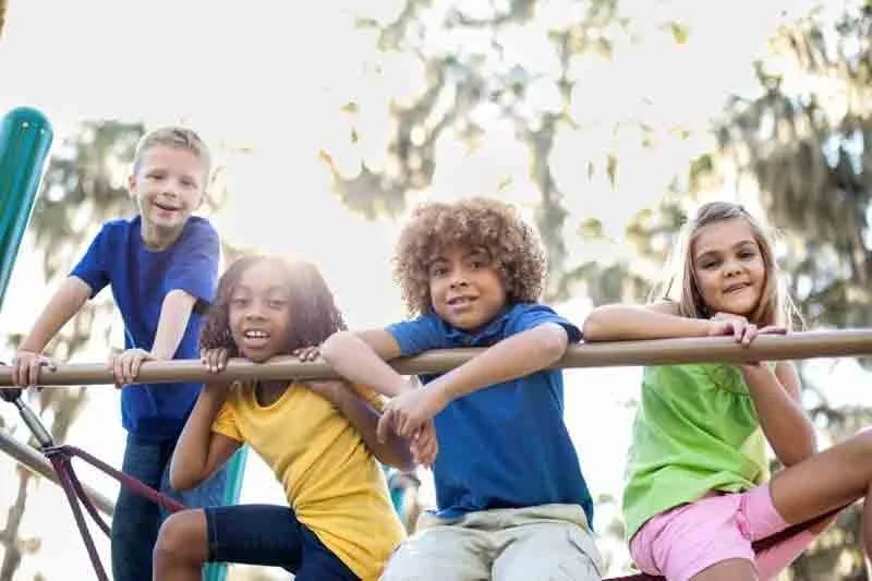 Four children smiling and holding onto a horizontal bar on a playground, with trees in the background.
