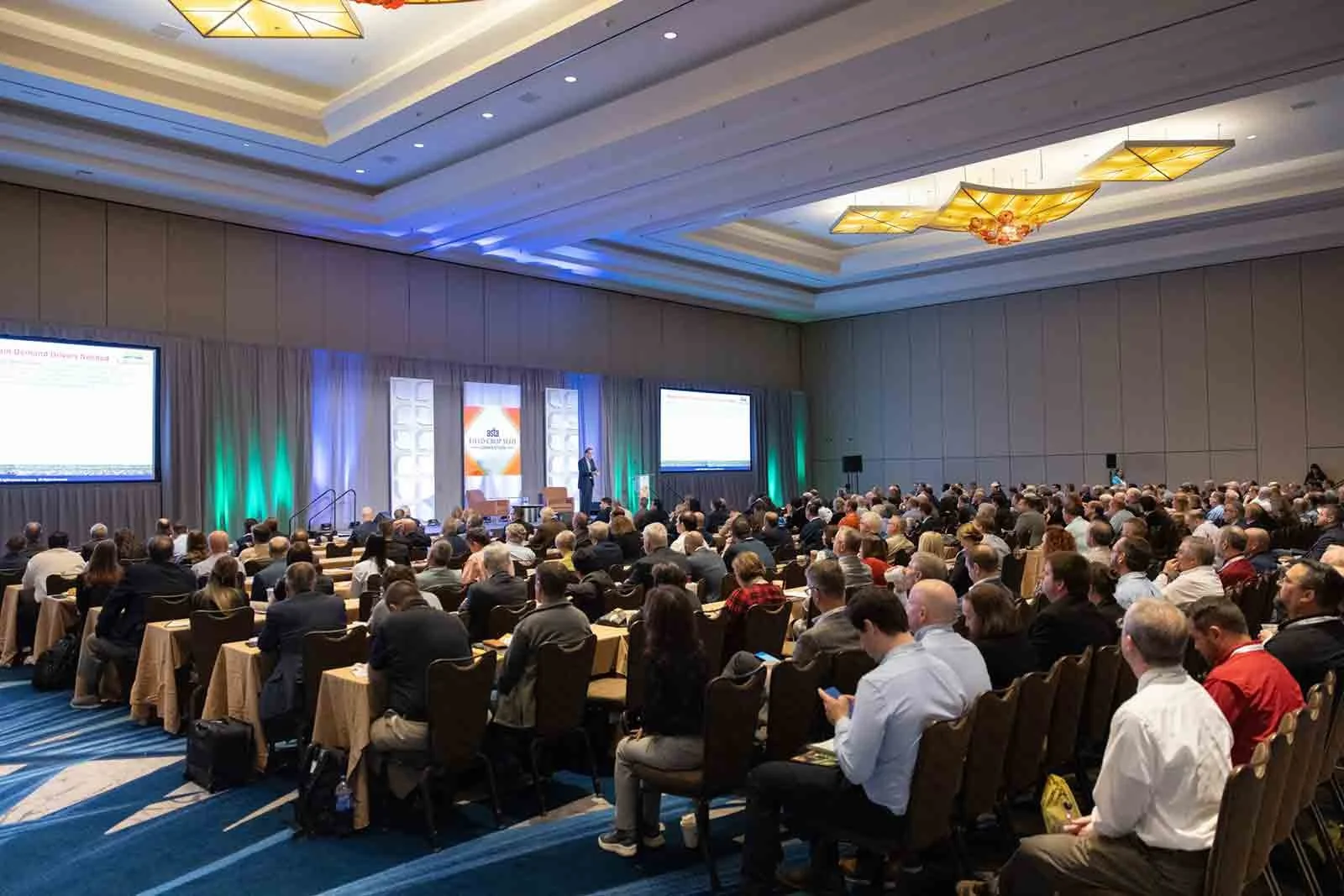 A large audience seated in rows at a conference, facing a stage with a speaker, large screens, and colorful lighting inside a spacious conference hall.