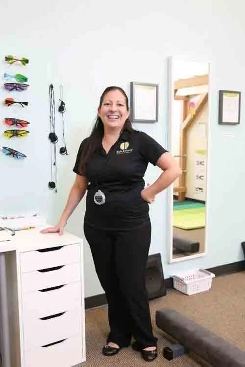 A smiling woman standing in a fitness or wellness room with equipment, sunglasses display, mirror, and a bunk bed in the background.