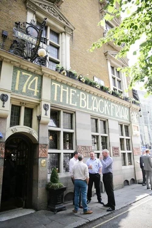Exterior of The Black Friar pub with a group of four men conversing outside on a sidewalk, building features traditional architecture with large windows, decorative trim, and a sign displaying the pub's name.
