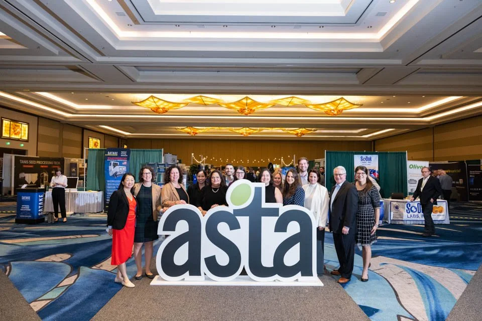 Group of conference attendees posing behind large 'asta' sign at a professional event in a hotel ballroom with booths in the background.