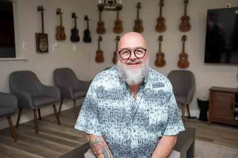 A smiling man with glasses and a beard, wearing a patterned shirt, sitting in a room with guitars hanging on the wall and gray chairs in the background.