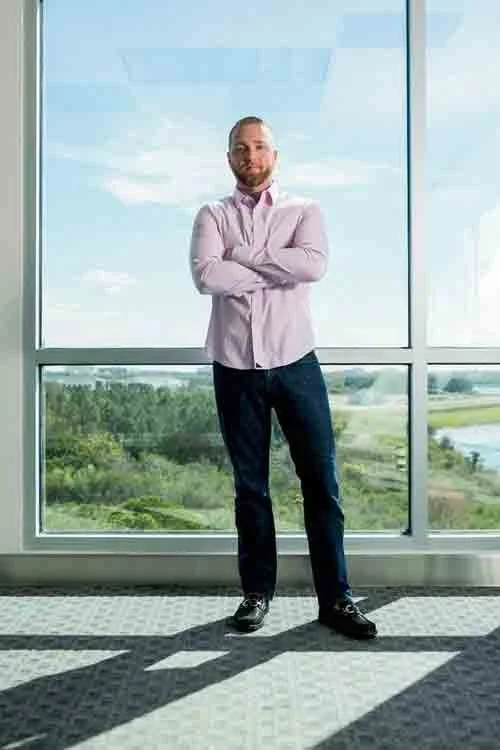 A man standing with arms crossed in front of a large window overlooking a green landscape and a river.