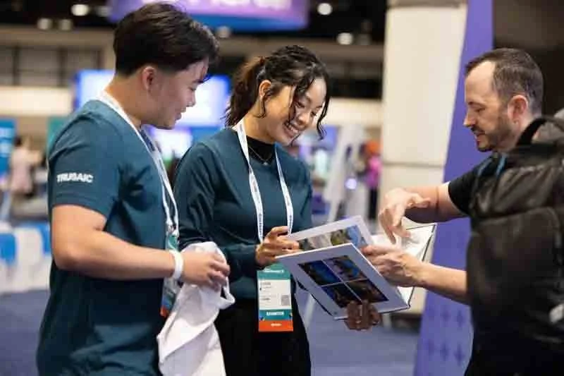 Two young women in teal shirts and a man in black shirt exchanging documents or brochures at a conference or expo.