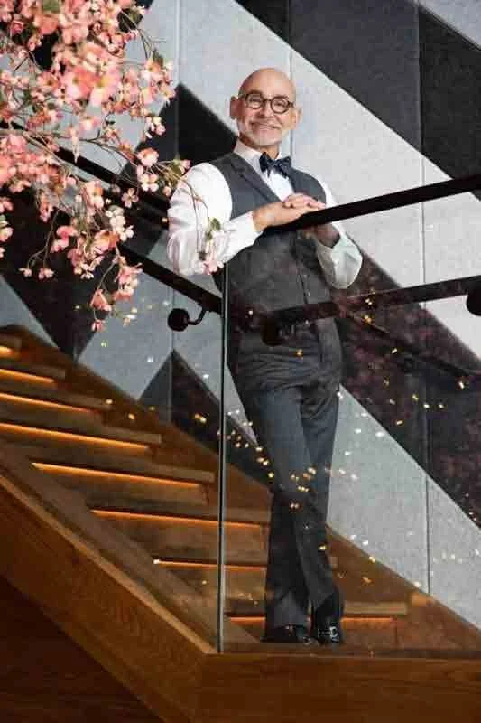 A man dressed in a tuxedo and glasses standing on a staircase with glass railing, holding his hands together, smiling, beside pink flowers and a modern interior.
